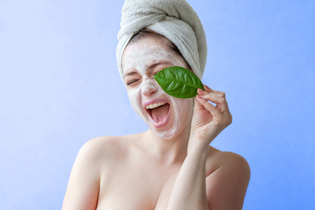 Beauty portrait of a smiling brunette woman in a towel on the head with white nourishing mask or creme on face and green leaf in hand on blue background isolated. Skincare cleansing eco organic cosmetic spa relax conceptの写真素材
