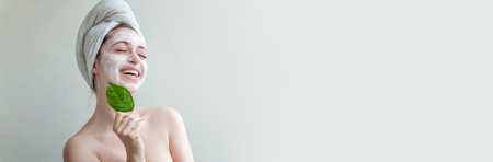 Beauty portrait of woman in towel on head with white nourishing mask or creme on face and green leaf in hand, white background isolated. Skincare cleansing eco organic cosmetic spa concept Bannerの写真素材