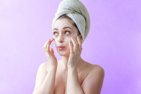 Beauty portrait of a smiling brunette woman in a towel on the head applying white nourishing mask or creme on face on purple background isolated. Skincare cleansing spa relax conceptの写真素材