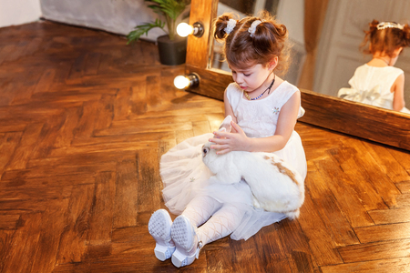 Easter celebration, pet care and animals concept. Cute happy little girl sitting on floor, holding and playing with adorable white fluffy rabbit bunny in modern room over mirrorの写真素材