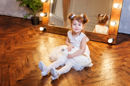 Easter celebration, pet care and animals concept. Cute happy little girl sitting on floor, holding and playing with adorable white fluffy rabbit bunny in modern room over mirrorの写真素材