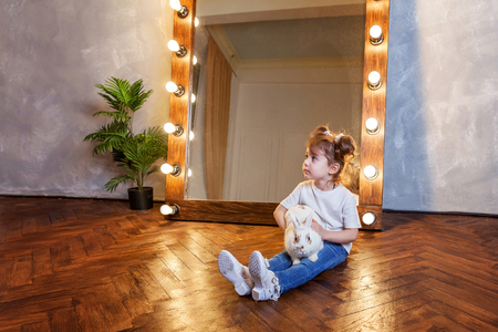 Easter celebration, pet care and animals concept. Cute happy little girl sitting on floor, holding and playing with adorable white fluffy rabbit bunny in modern room over mirrorの写真素材