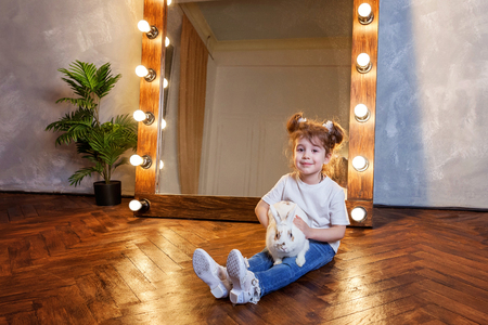 Easter celebration, pet care and animals concept. Cute happy little girl sitting on floor, holding and playing with adorable white fluffy rabbit bunny in modern room over mirrorの写真素材