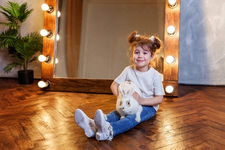 Easter celebration, pet care and animals concept. Cute happy little girl sitting on floor, holding and playing with adorable white fluffy rabbit bunny in modern room over mirrorの写真素材