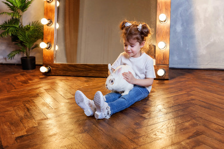Easter celebration, pet care and animals concept. Cute happy little girl sitting on floor, holding and playing with adorable white fluffy rabbit bunny in modern room over mirrorの写真素材