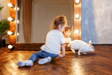 Easter celebration, pet care and animals concept. Cute happy little girl sitting on floor, holding and playing with adorable white fluffy rabbit bunny in modern room over mirrorの写真素材
