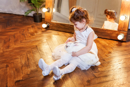 Easter celebration, pet care and animals concept. Cute happy little girl sitting on floor, holding and playing with adorable white fluffy rabbit bunny in modern room over mirrorの写真素材