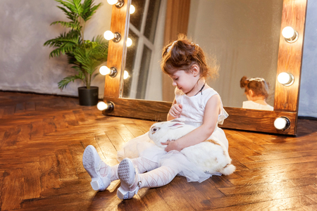 Easter celebration, pet care and animals concept. Cute happy little girl sitting on floor, holding and playing with adorable white fluffy rabbit bunny in modern room over mirrorの写真素材