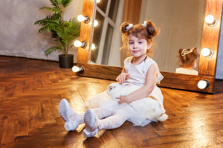 Easter celebration, pet care and animals concept. Cute happy little girl sitting on floor, holding and playing with adorable white fluffy rabbit bunny in modern room over mirrorの写真素材