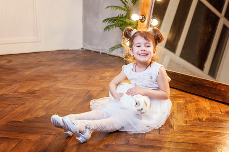 Easter celebration, pet care and animals concept. Cute happy little girl sitting on floor, holding and playing with adorable white fluffy rabbit bunny in modern room over mirrorの写真素材