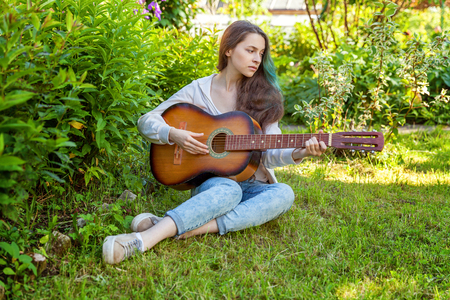 Young hipster woman sitting in grass and playing guitar on park or garden background. Teen girl learning to play song and writing music. Hobby, lifestyle, relax, Instrument, leisure, education conceptの写真素材