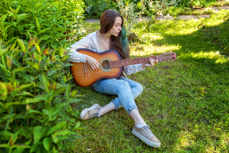 Young hipster woman sitting in grass and playing guitar on park or garden background. Teen girl learning to play song and writing music. Hobby, lifestyle, relax, Instrument, leisure, education conceptの写真素材