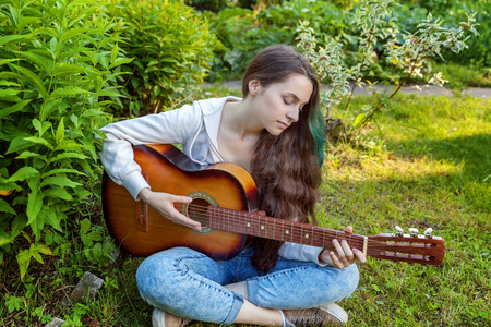 Young hipster woman sitting in grass and playing guitar on park or garden background. Teen girl learning to play song and writing music. Hobby, lifestyle, relax, Instrument, leisure, education conceptの写真素材