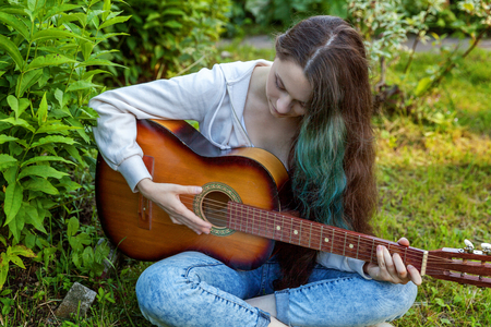 Young hipster woman sitting in grass and playing guitar on park or garden background. Teen girl learning to play song and writing music. Hobby, lifestyle, relax, Instrument, leisure, education conceptの写真素材