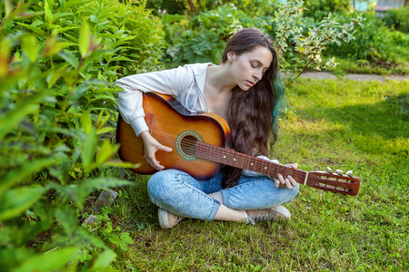 Young hipster woman sitting in grass and playing guitar on park or garden background. Teen girl learning to play song and writing music. Hobby, lifestyle, relax, Instrument, leisure, education conceptの写真素材