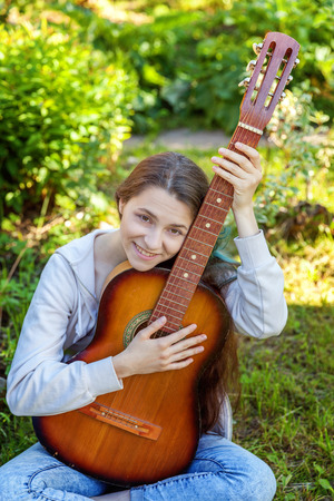 Young hipster woman sitting in grass and playing guitar on park or garden background. Teen girl learning to play song and writing music. Hobby, lifestyle, relax, Instrument, leisure, education conceptの写真素材