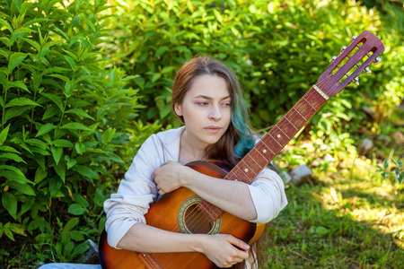 Young hipster woman sitting in grass and playing guitar on park or garden background. Teen girl learning to play song and writing music. Hobby, lifestyle, relax, Instrument, leisure, education conceptの写真素材