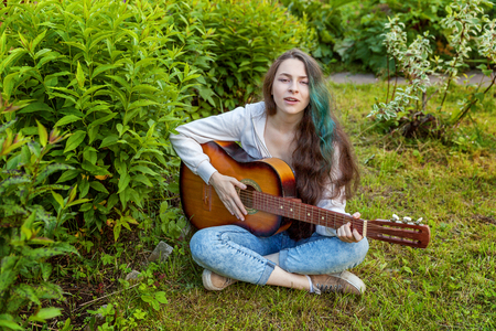 Young hipster woman sitting in grass and playing guitar on park or garden background. Teen girl learning to play song and writing music. Hobby, lifestyle, relax, Instrument, leisure, education conceptの写真素材