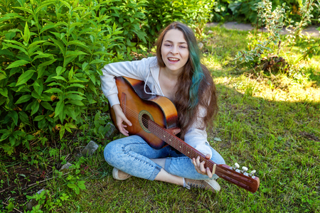 Young hipster woman sitting in grass and playing guitar on park or garden background. Teen girl learning to play song and writing music. Hobby, lifestyle, relax, Instrument, leisure, education conceptの写真素材