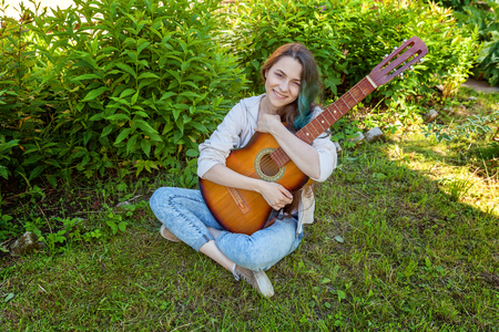 Young hipster woman sitting in grass and playing guitar on park or garden background. Teen girl learning to play song and writing music. Hobby, lifestyle, relax, Instrument, leisure, education conceptの写真素材
