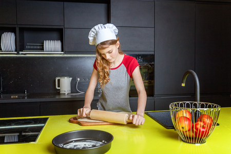 Teenage girl with chef hat, rolling pin preparing dough, bake homemade holiday apple pie in kitchen. Young woman cooking healthy food at home and having fun. Food, healthy eating, household conceptの写真素材