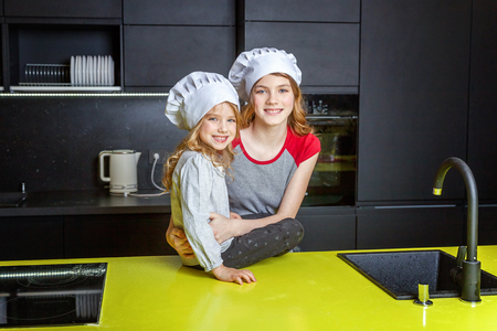 Two girls with chef hat hugging and having fun in kitchen. Sisters little kid and teenage girl cooking healthy food at home. Childhood, family, household, teamwork helping conceptの写真素材