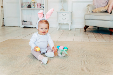Little child girl wearing bunny ears on Easter day. Girl holding basket with painted eggs sitting on floor at home, having fun on Easter egg hunt. Happy Easter holiday celebration spring conceptの写真素材