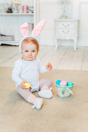 Little child girl wearing bunny ears on Easter day. Girl holding basket with painted eggs sitting on floor at home, having fun on Easter egg hunt. Happy Easter holiday celebration spring conceptの写真素材