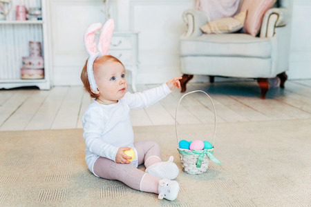 Little child girl wearing bunny ears on Easter day. Girl holding basket with painted eggs sitting on floor at home, having fun on Easter egg hunt. Happy Easter holiday celebration spring conceptの写真素材