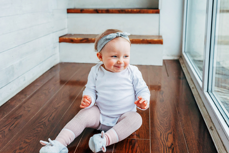 Little crawling baby girl one year old siting on floor in bright light living room near window smiling and laughing. Happy toddler kid playing at home. Childhood conceptの写真素材