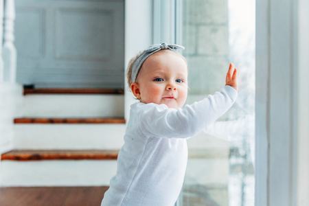 Little crawling baby girl one year old siting on floor in bright light living room near window...