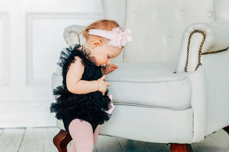Little baby girl one year old wearing spring wreath standing near cozy chair in bright light living...