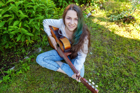 Young hipster woman sitting in grass and playing guitar on park or garden background. Teen girl learning to play song and writing music. Hobby, lifestyle, relax, Instrument, leisure, education conceptの写真素材