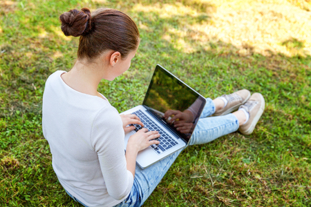 Freelance business concept. Young woman sitting on green grass lawn in city park working on laptop pc computer. Lifestyle authentic candid student girl studying outdoors. Mobile Officeの写真素材