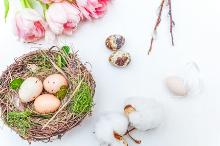 Spring greeting card. Easter eggs in nest cotton willow branch and pink fresh tulip flowers bouquet on rustic white wooden background. Easter concept. Flat lay top view copy space. Spring flowers tulipsの写真素材