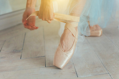 Hands of ballerina in blue tutu skirt puts on pointe shoes on leg in white light hall. Young...