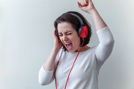 Energy young brunette lady woman listening music in headphones and singing isolated on white background. Charming caucasian girl in earphones enjoying music and dancing with closed eyesの写真素材