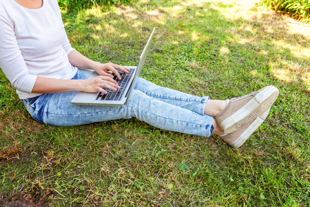 Freelance business concept. Woman legs on green grass lawn in city park, hands working on laptop pc computer. Lifestyle authentic candid student girl studying outdoors. Mobile Officeの写真素材