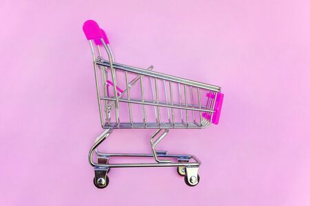 Small supermarket grocery push cart for shopping toy with wheels and pink plastic elements on pink pastel color paper flat lay background. Concept of shopping. Copy space for advertisementの写真素材