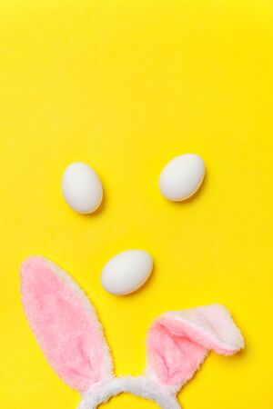 Happy Easter concept. Preparation for holiday. Decorative white eggs and bunny ears furry costume toy isolated on trendy yellow background. Simple minimalism flat lay top view copy spaceの写真素材
