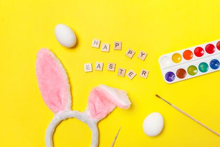 Happy Easter concept. Preparation for holiday. Inscription HAPPY EASTER letters eggs colorful paints and bunny ears isolated on trendy yellow background. Simple minimalism flat lay top view copy spaceの写真素材