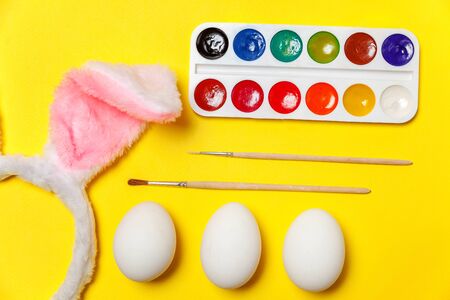 Happy Easter concept. Preparation for holiday. Decorative eggs colorful paints and bunny ears furry costume toy isolated on trendy yellow background. Simple minimalism flat lay top view copy spaceの写真素材