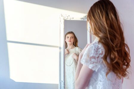 beauty, hygiene, morning and people concept - smiling happy teenage girl looking at reflection in mirror. young positive woman wearing white dress posing in bright light room against white wallの写真素材