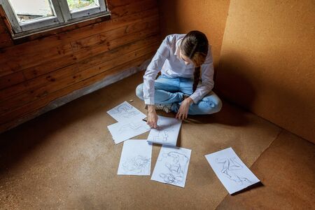 Young hipster woman sitting on floor and drawing sketch in album at home. Teen girl learning...