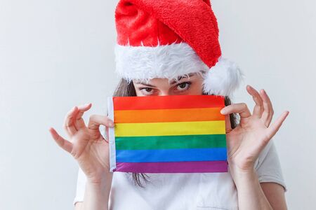 Beautiful lesbian girl in red Santa Claus hat with LGBT rainbow flag isolated on white background looking happy and excited. Young woman Gay Pride portrait. Happy Christmas and New Year holidays.の写真素材