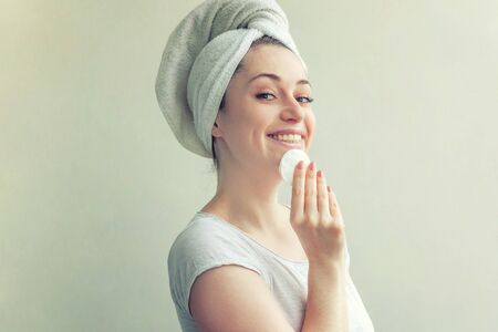 Beauty portrait of smiling woman in towel on head with soft healthy skin removing make up with cotton pad isolated on white background. Skincare cleansing spa relax conceptの写真素材