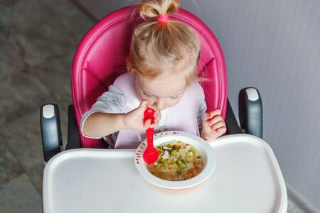 Infant baby with dirty face eating soup herself with spoon. Cute little girl sitting in high baby chair in kitchen at home. Maternity family childhood conceptの写真素材