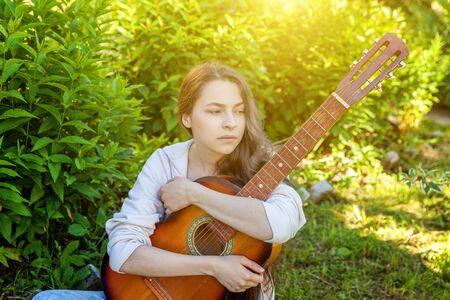 Young hipster woman sitting in grass and playing guitar on park or garden background. Teen girl...