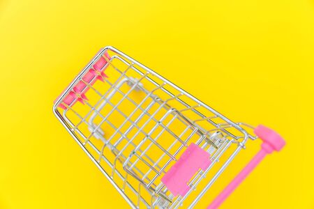 Small supermarket grocery push cart for shopping toy with wheels isolated on yellow colourful trendy modern fashion background. Sale buy mall market shop consumer concept. Copy spaceの写真素材