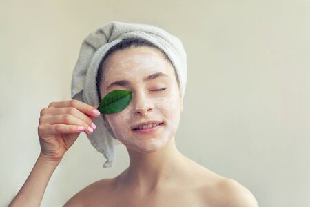 Beauty portrait of woman in towel on head with white nourishing mask or creme on face and green leaf in hand, white background isolated. Skincare cleansing eco organic cosmetic spa conceptの写真素材